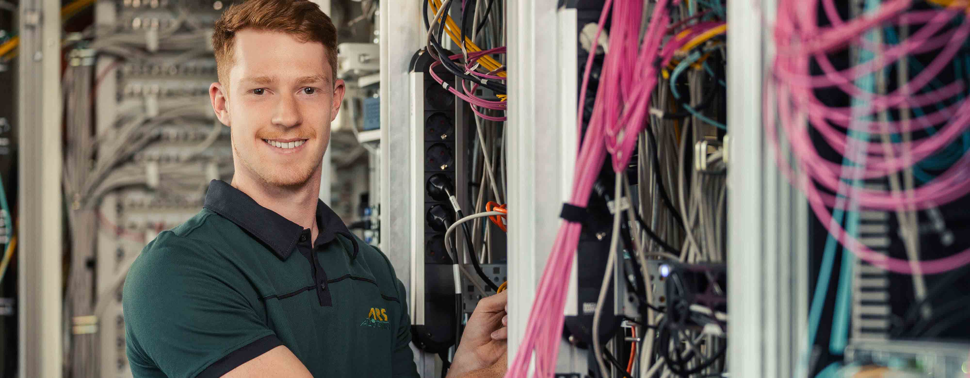 Person, die in einem Serverraum neben Kabelschränken steht und in die Kamera lächelt.