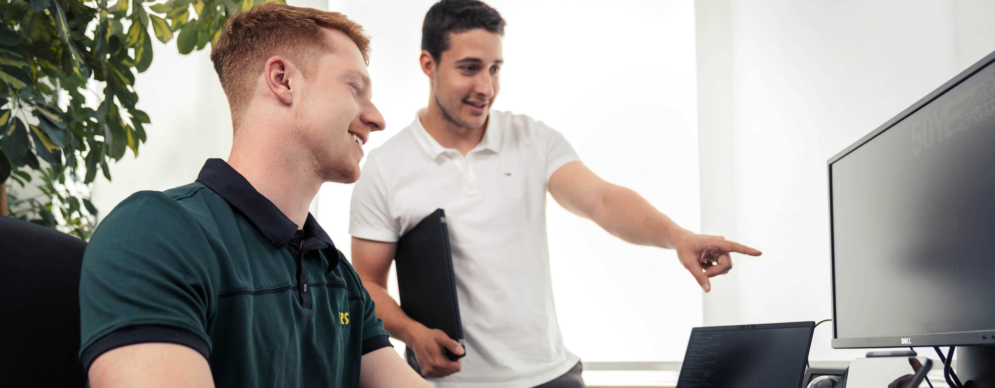 Zwei Personen, die an einem PC sitzen. Die eine Person zeigt mit dem Finger auf den Bildschirm.