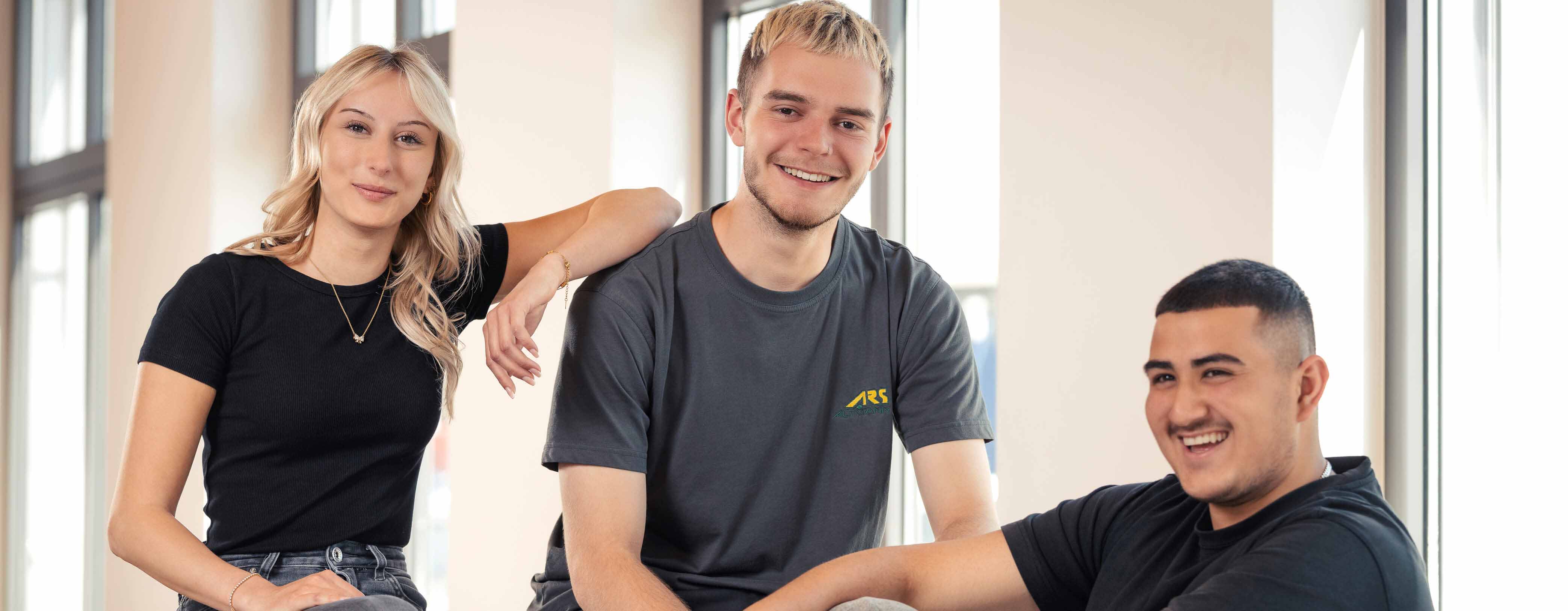 Drei Junge Personen, die locker nebeneinander sitzen und in die Kamera lächeln.